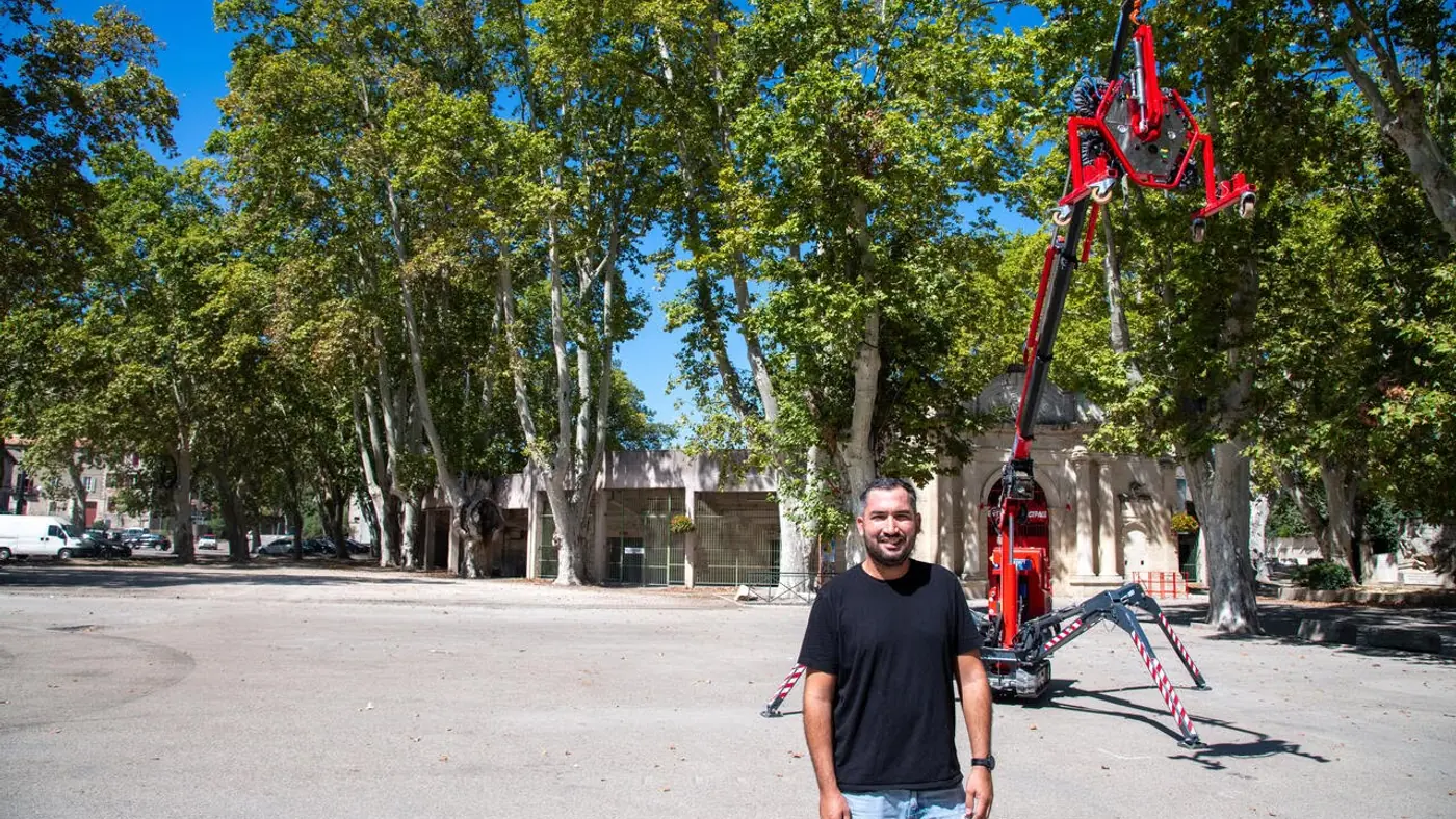 Joris Cervantes debout à côté de la mini-grue araignée repliée, sous une allée de platanes