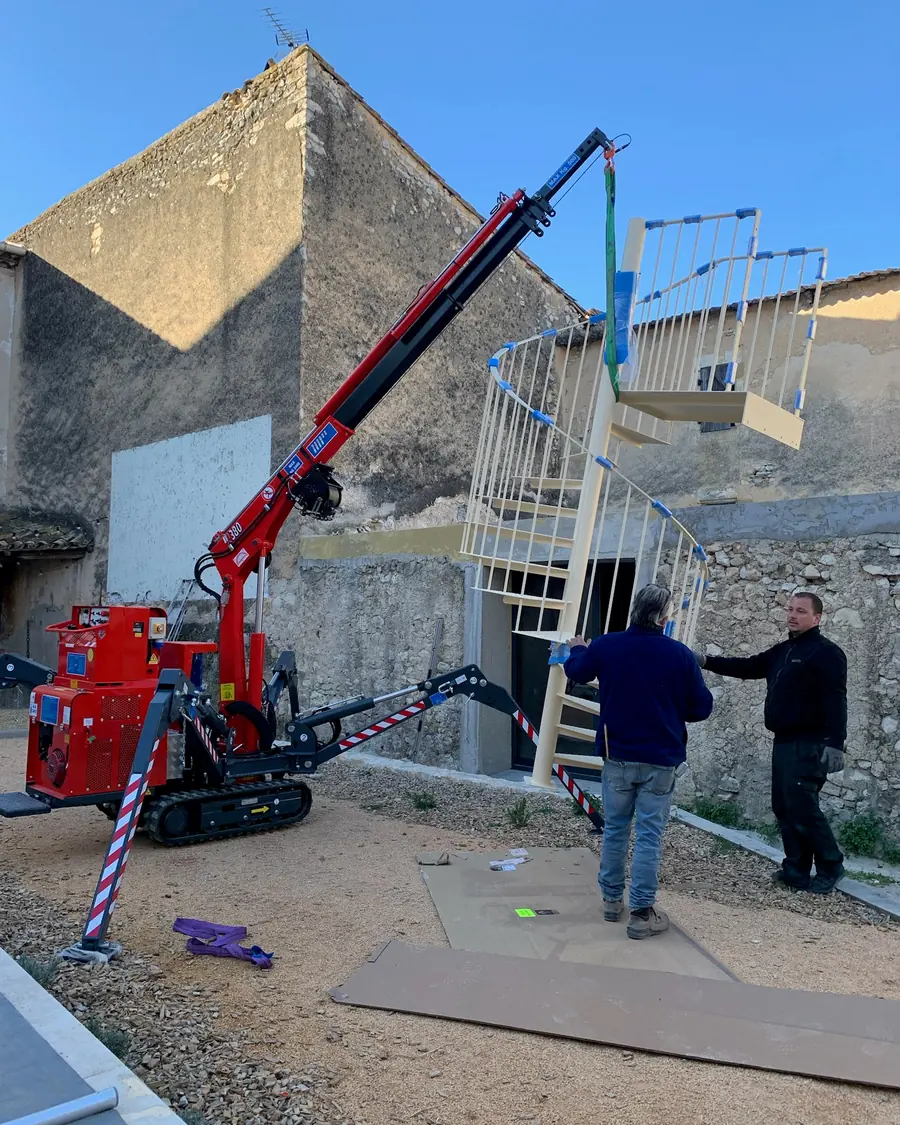 Mini-grue araignée présentant un escalier métallique en colimaçon dans une cour, deux personnes le guidant